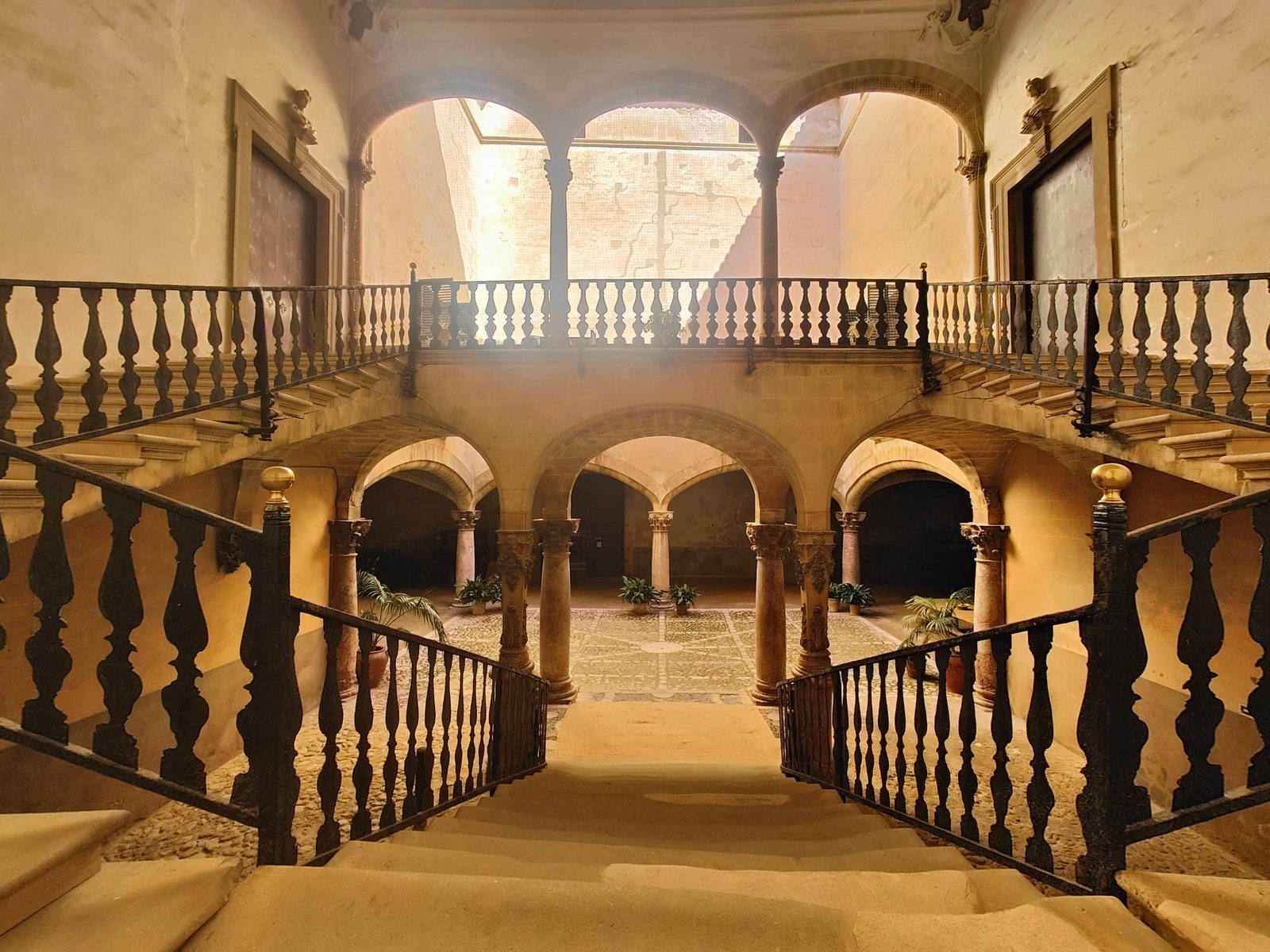 The courtyard of Can Vivot seen from the top of its stone staircase: two tiers of arches on marble columns, a balustrade, and afternoon light coming down through the open roof.