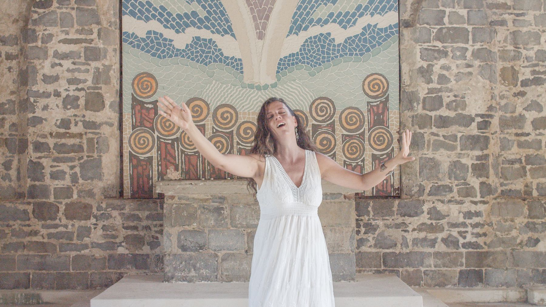 Cumie singing with her arms open wide in a white dress, in a stone chapel in front of a large mosaic.